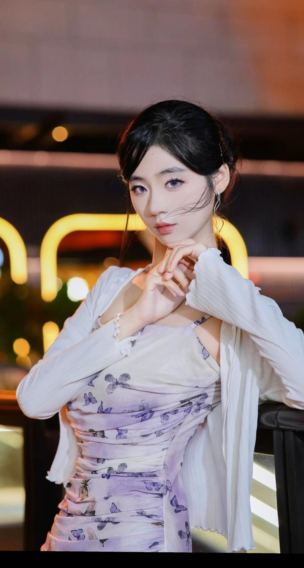 一位身着淡紫色蝴蝶印花吊带连衣裙搭配白色薄纱开衫的女性，在夜晚城市灯光背景下的多