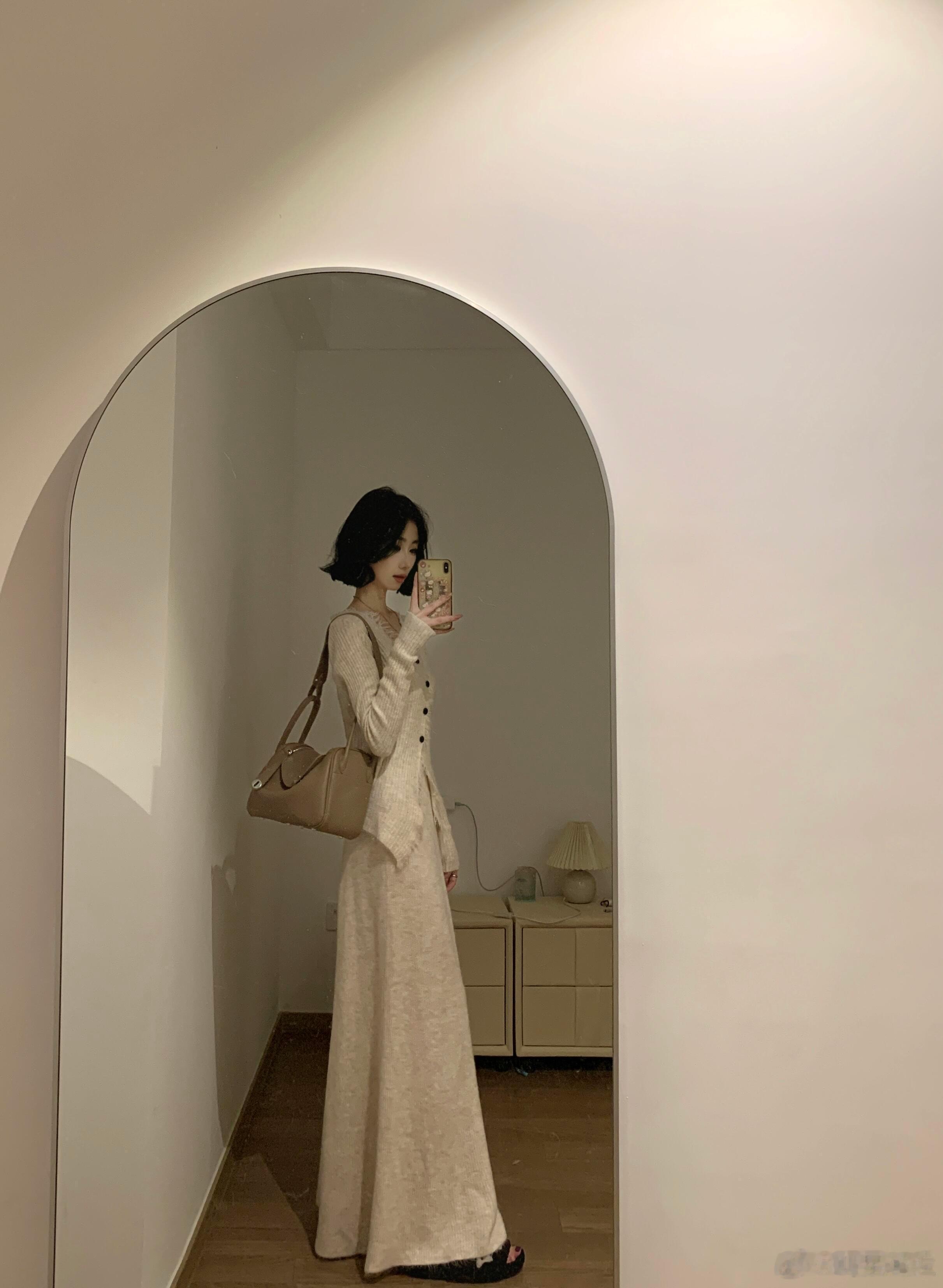 图中女子在拱形镜子前自拍。她留着黑色短发，身着米白色针织套装，上装为V领开衫，下