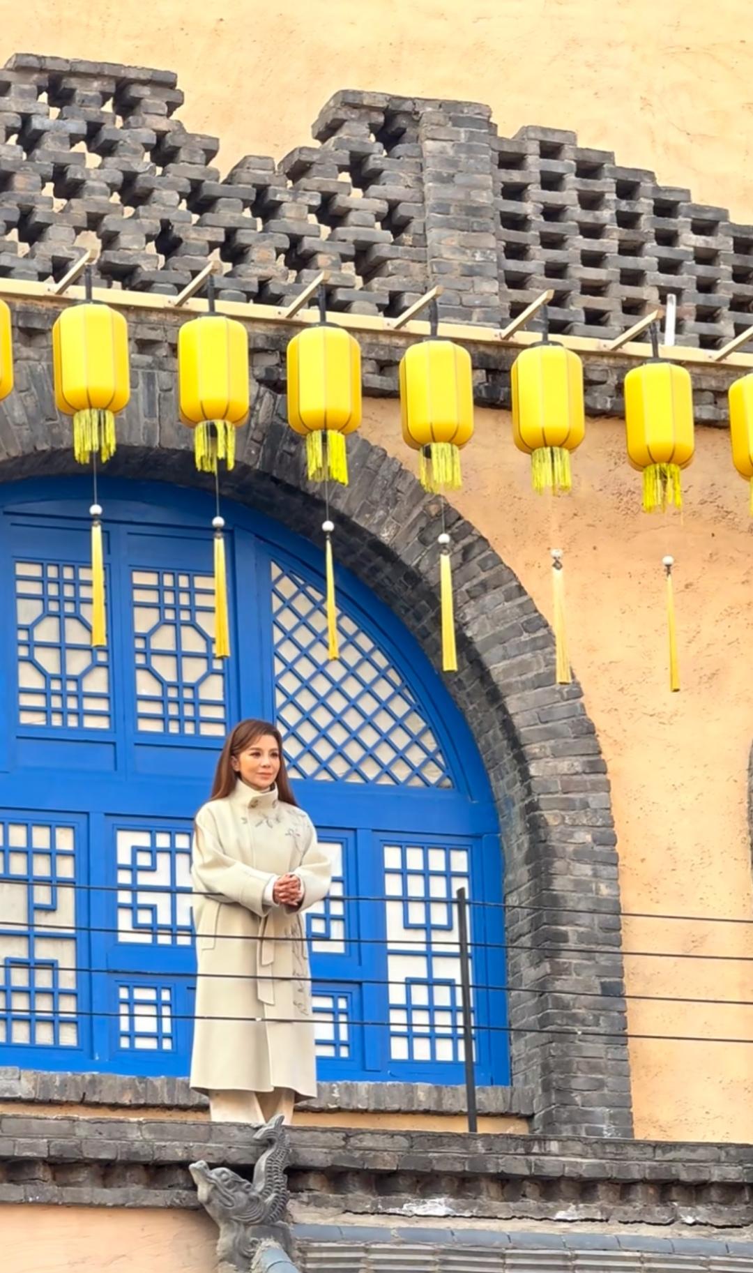 1月6日，朱迅打卡河南戏剧幻城，身着暖白色立领大衣，衣身纹样雅致，安静地立于青砖