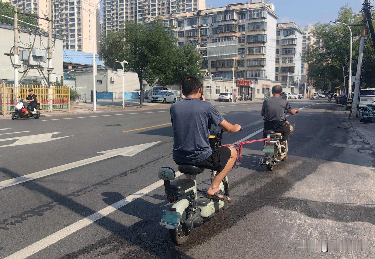前方一辆电动车在前行驶，通过一根红色绳索牵引着后方另一辆电动车，两车在道路上同向