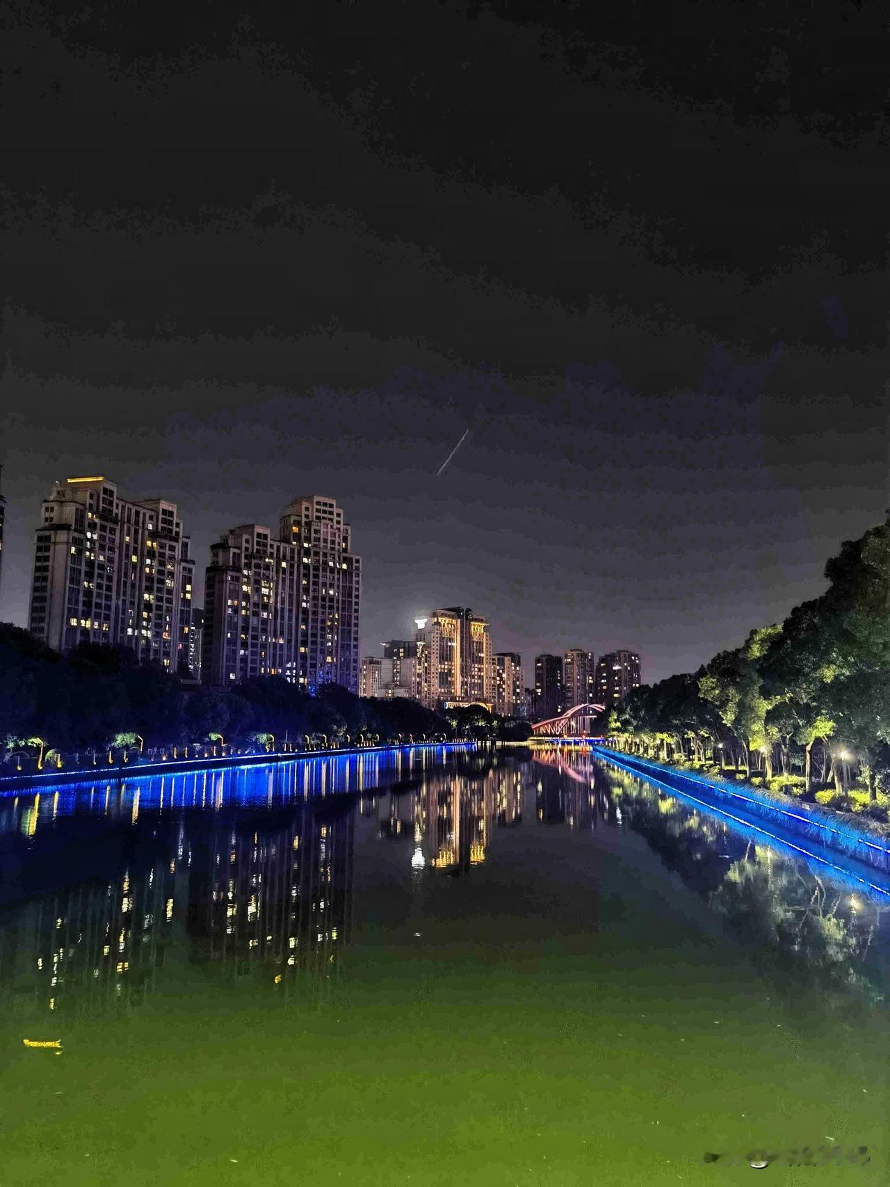 夜色沉静，水波如镜，高楼暖光与沿岸的蓝色灯带交相辉映，倒影在水面拉出长长画卷。