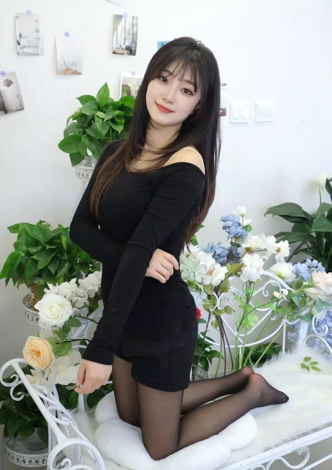 她穿着黑色修身连衣裙和黑丝袜，跪坐在浅灰色沙发上。背景是贴着软木板的白墙，整体氛