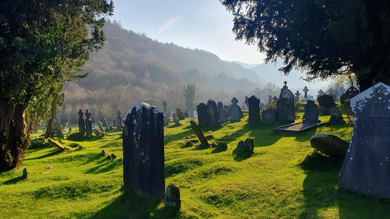 这是爱尔兰威克洛郡格伦达洛的一处古老墓地，画面中可见一座圆形石塔和散落的十字墓碑