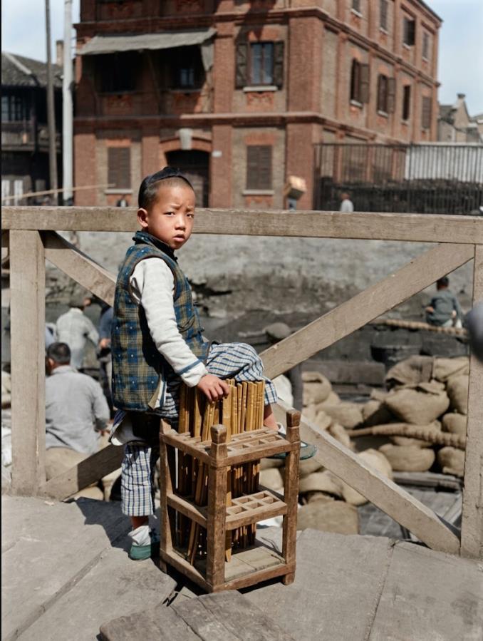 1917年，南京街头，一个保留小辫的幼童，穿格纹马甲与短裤，站在木栏杆旁，手扶一