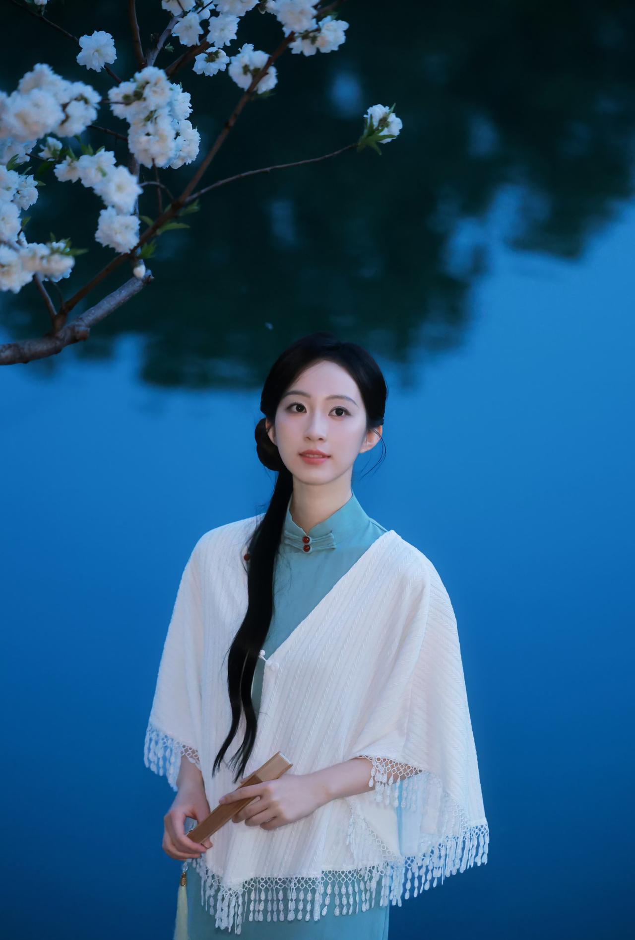 暮色浸湖蓝，花枝缀雪白，
一袭青衫配素色披肩，手执轻扇，
眉目温婉，似从江南旧梦