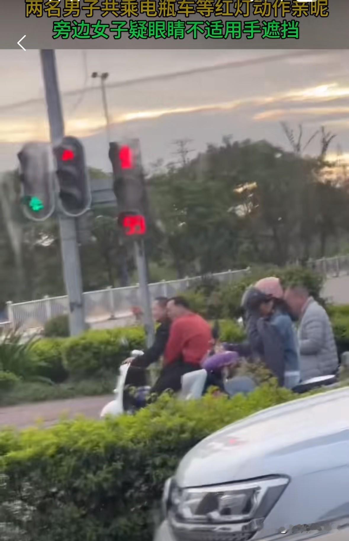 两个中年男子同骑一辆电瓶车，而且举止非常亲密。
后面的红衣男子紧紧的抱着骑车的男