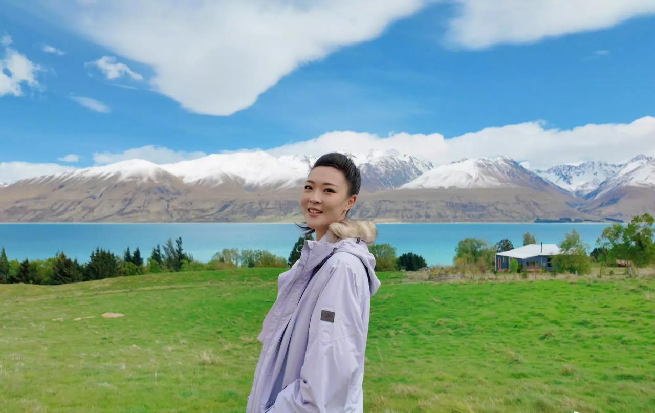 远处的蓝天白云，天空下面是白色巍峨的雪山。雪山下面蓝色的湖水。她站在湖边的绿色草