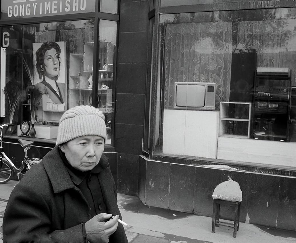 80年代的老成都街头，一位手拿香烟的男子从商店门口路过，商店的橱窗里还摆放着电视