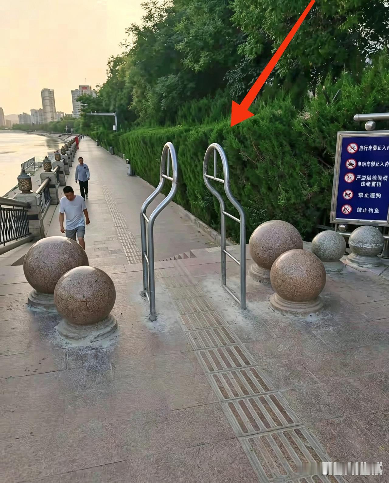某城市滨河步行道入口，一旁是河道护栏、休闲步道，路边布置绿化与隔离石球，边上还立