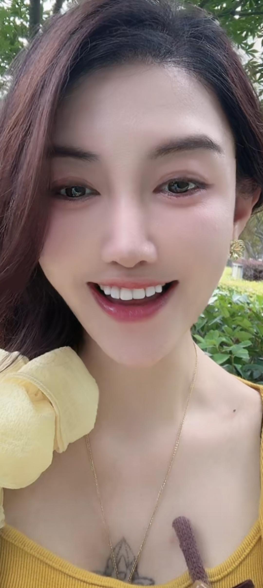 笑容灿烂，穿着黄色上衣，背景是绿树。