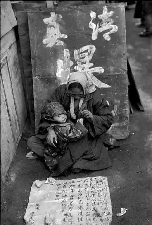 1948年12月，北平城内乞讨的母子。
裹着头巾、身着破旧衣服的妇人，怀抱着年幼