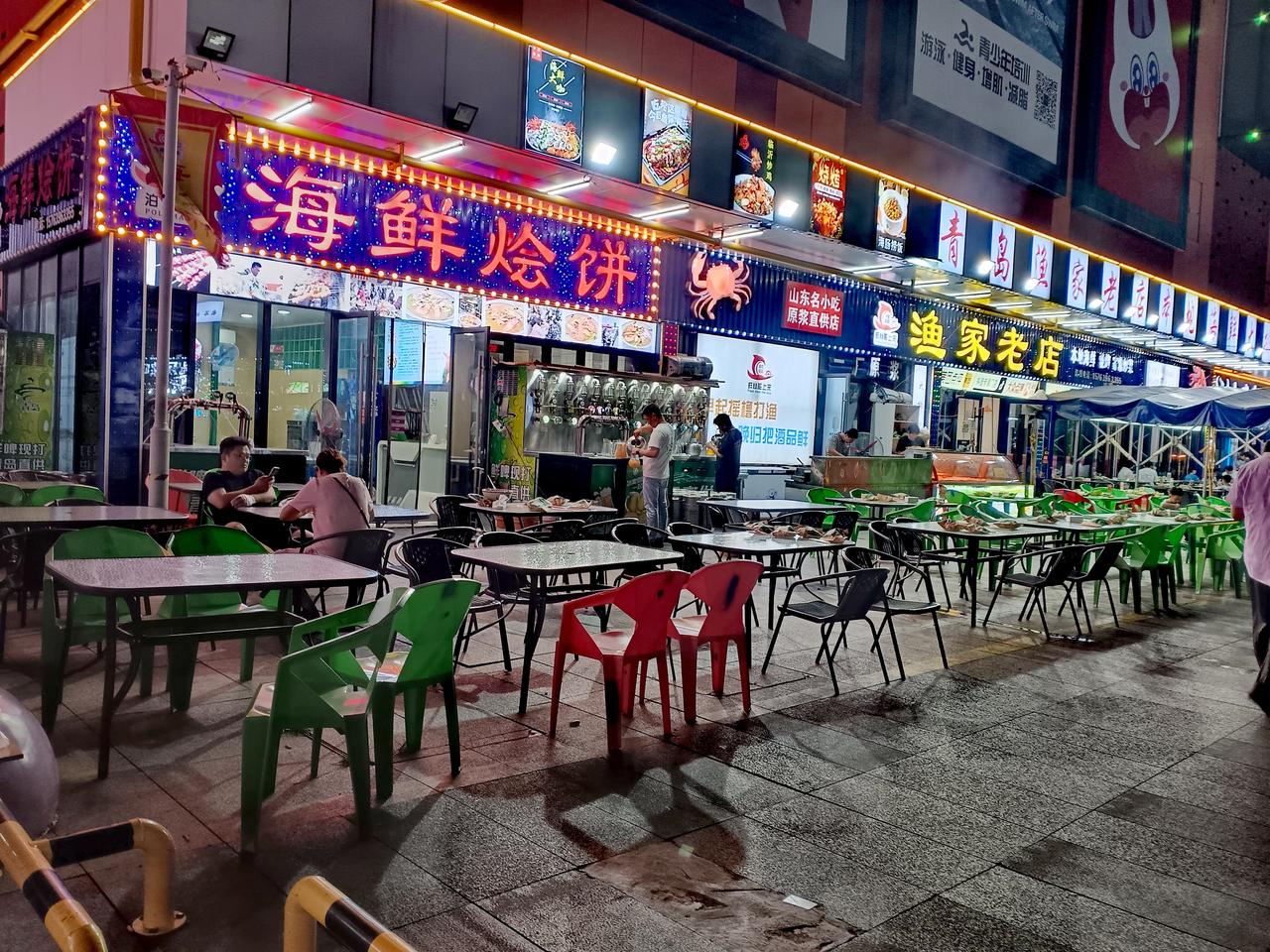 霓虹招牌点亮海鲜夜市，桌椅沿街排布，海风裹挟烟火气，满是滨海夜宵的热闹惬意。