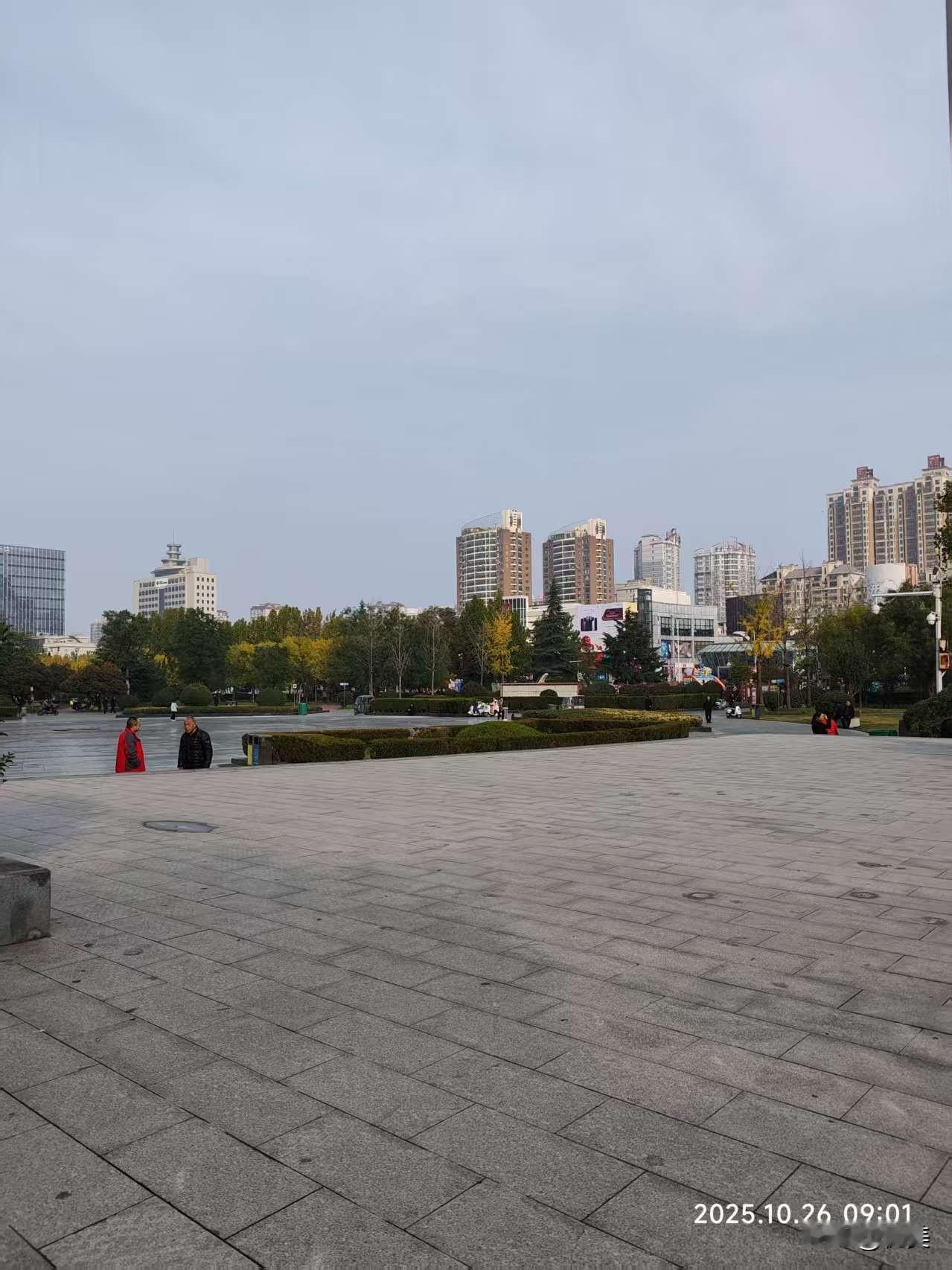 清晨的广场开阔明净，灰石铺就的地面整洁舒展。远处高楼错落，秋树间杂黄绿，行人闲立