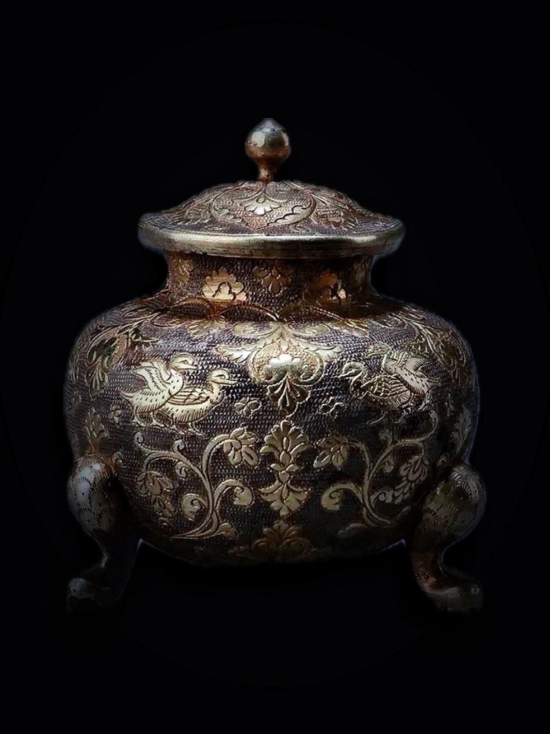 唐代 · 花鸟纹鎏金三足银罐。钟鼓馔玉不足贵，但愿长醉不复醒——唐·李白《将进酒》 
1989年西安市东郊国棉五厂65号唐墓出土，通高5.1厘米、腹径4.7厘米，陕西唐三彩艺术博物馆藏 
这件银罐小直口、短颈、广肩、鼓腹、圜底，中腹部附加三只蹄状足
带盖，盖覆杯形，盖顶有蘑菇形小钮 
罐体皆以密布的珠点纹为地纹，并被三组由四个心形团花图案组成的纹饰组分为三大块
每块以一足为中心，足上枯树枝上落有一对鸳鸯，前者顾首回望，后者展翅欲飞，生动形象，可爱逼真
鸳鸯周围有众多的折枝花纹作衬托，更具“绿叶红花”的效果