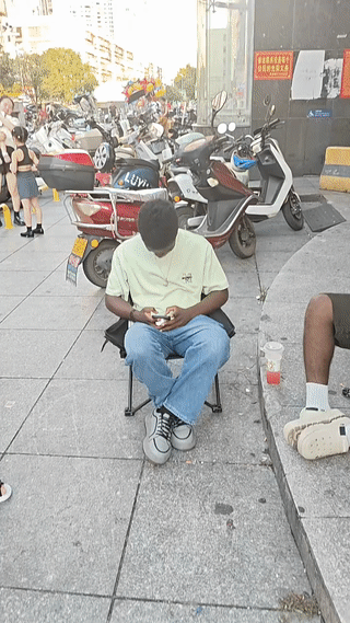 两名黑人男子坐在商场门口玩手机打游戏。
旁边的好像是还有人给他们俩人扇扇子？
这