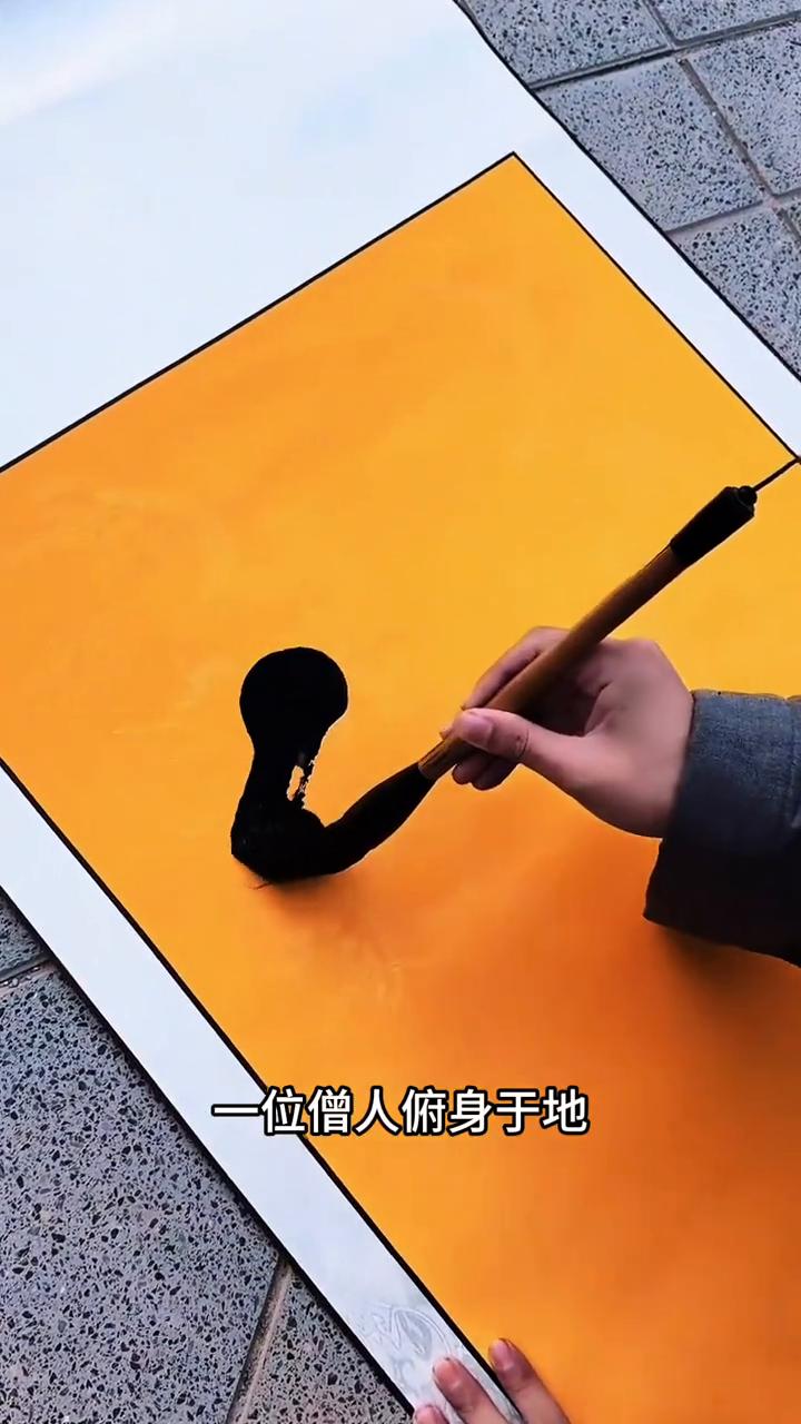 为僧人俯身于地，手握硕大毛笔，在黄纸上挥毫，先于左侧勾勒出一个小人，随即转向右侧