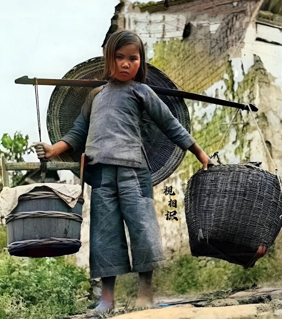 1944年，云南，一位挑着担子的小女孩的老照片，镜头中她赤着脚，穿着青布小褂和裤...
