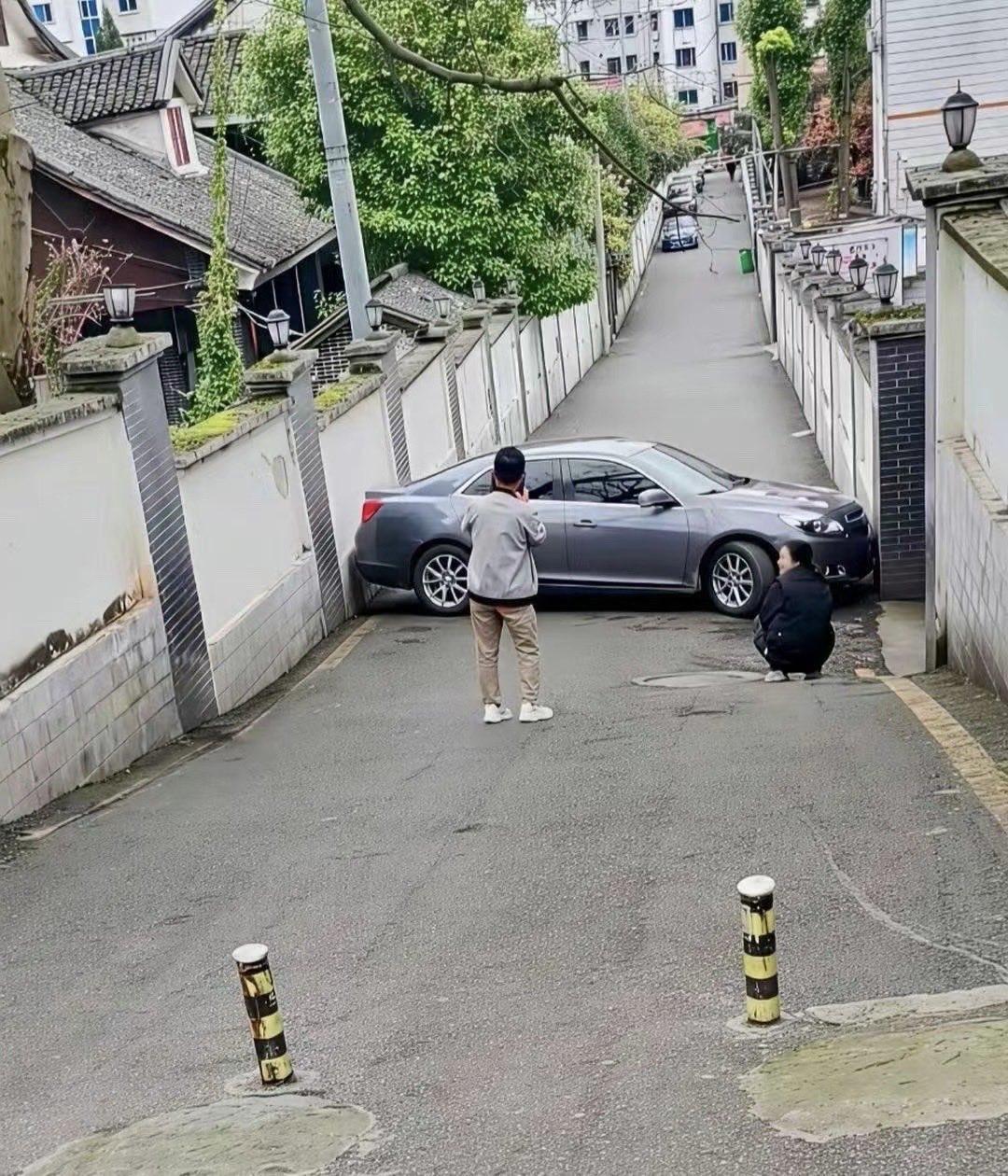 一辆灰色轿车尴尬地垂直停在一条狭窄的居民区巷子里，两名男子在一旁围观——一人站着