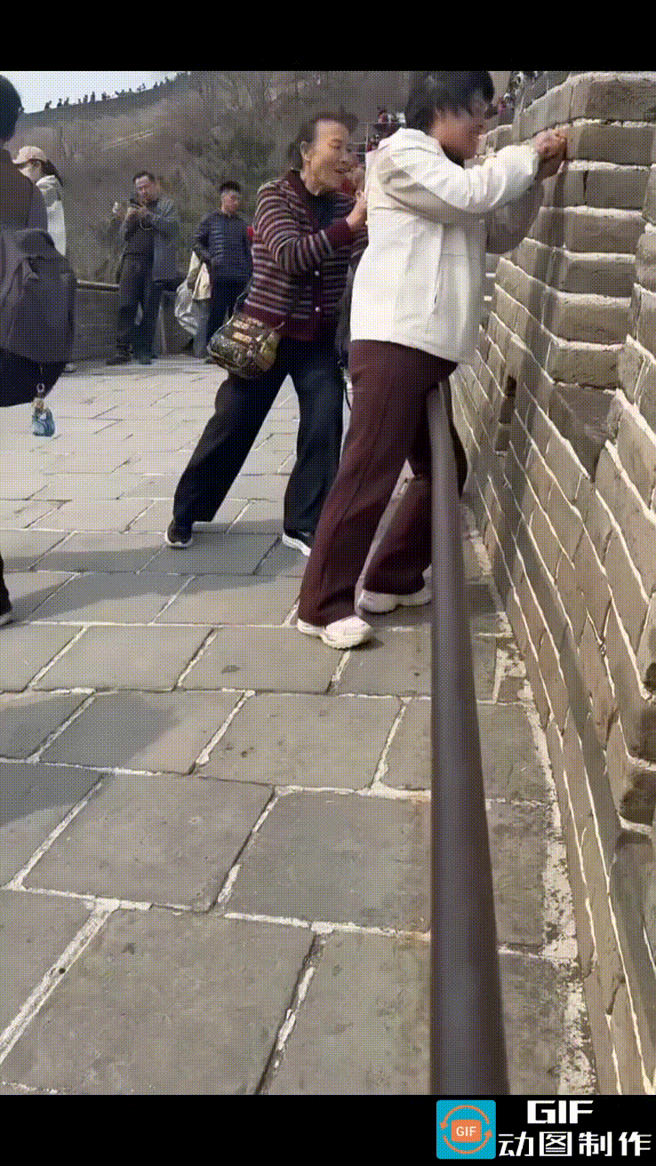 两名女子在八达岭长城墙体上刻字，一人拿尖锐的小石头用力在墙上划着，另一人举着手机