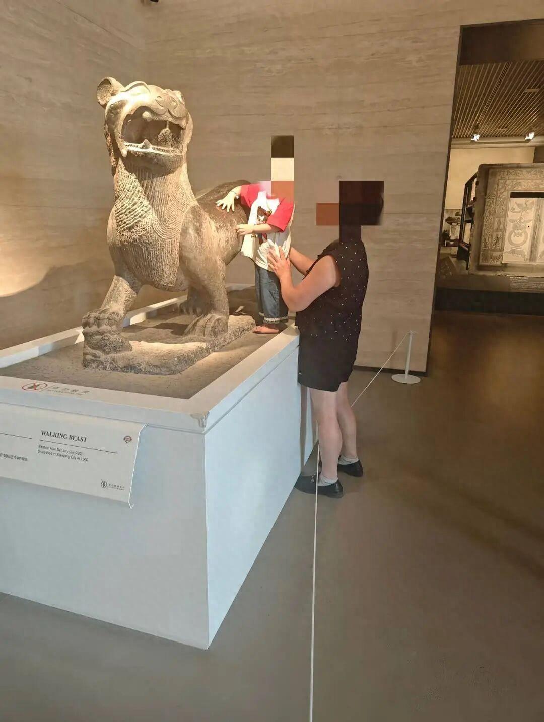 一个小孩，光脚站在博物馆的白色展台上，两只手直接摸在一尊石兽的背上。旁边一个大人