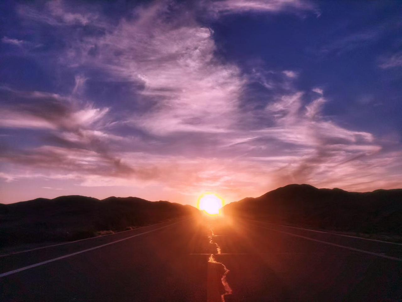 笔直公路向着群山延伸，落日悬在道路尽头，金辉铺满路面。漫天云霞晕开粉紫与橘红，远