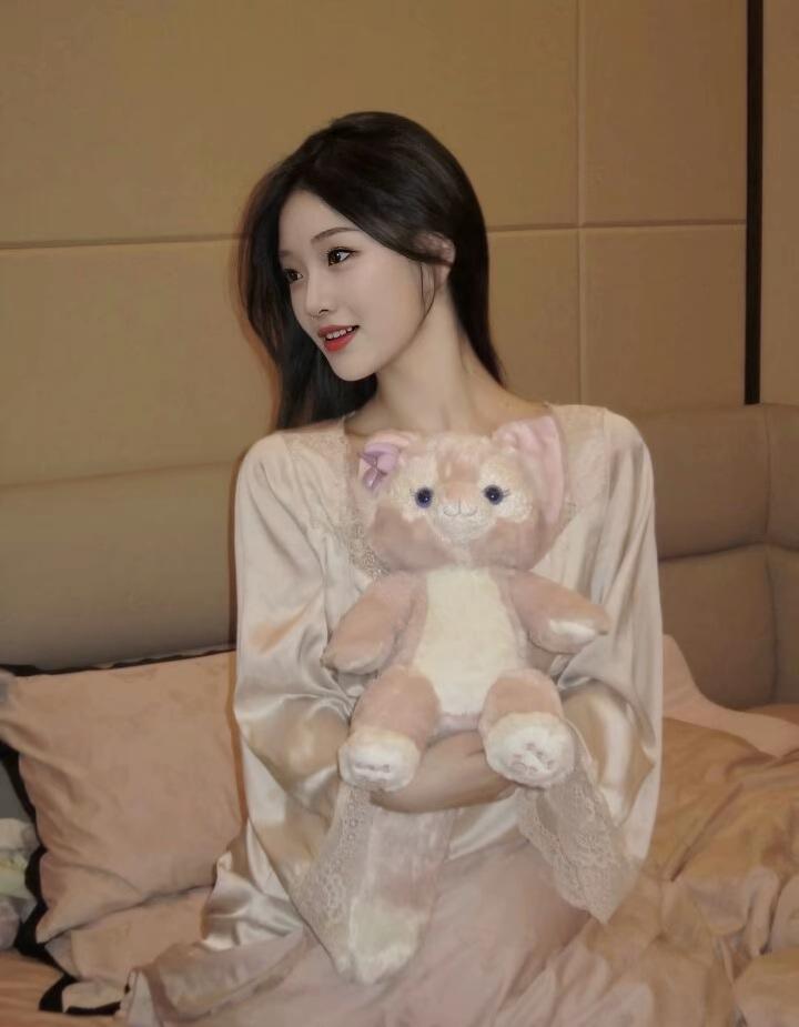 居家的她，身着浅粉色真丝蕾丝睡衣，慵懒地陷在柔软的床榻里。乌黑的长卷发随意披散，