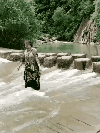 大妈来到景区游玩，她瞧见一个小型水库，顿时兴致勃勃地朝着水库下游走去，准备拍照留