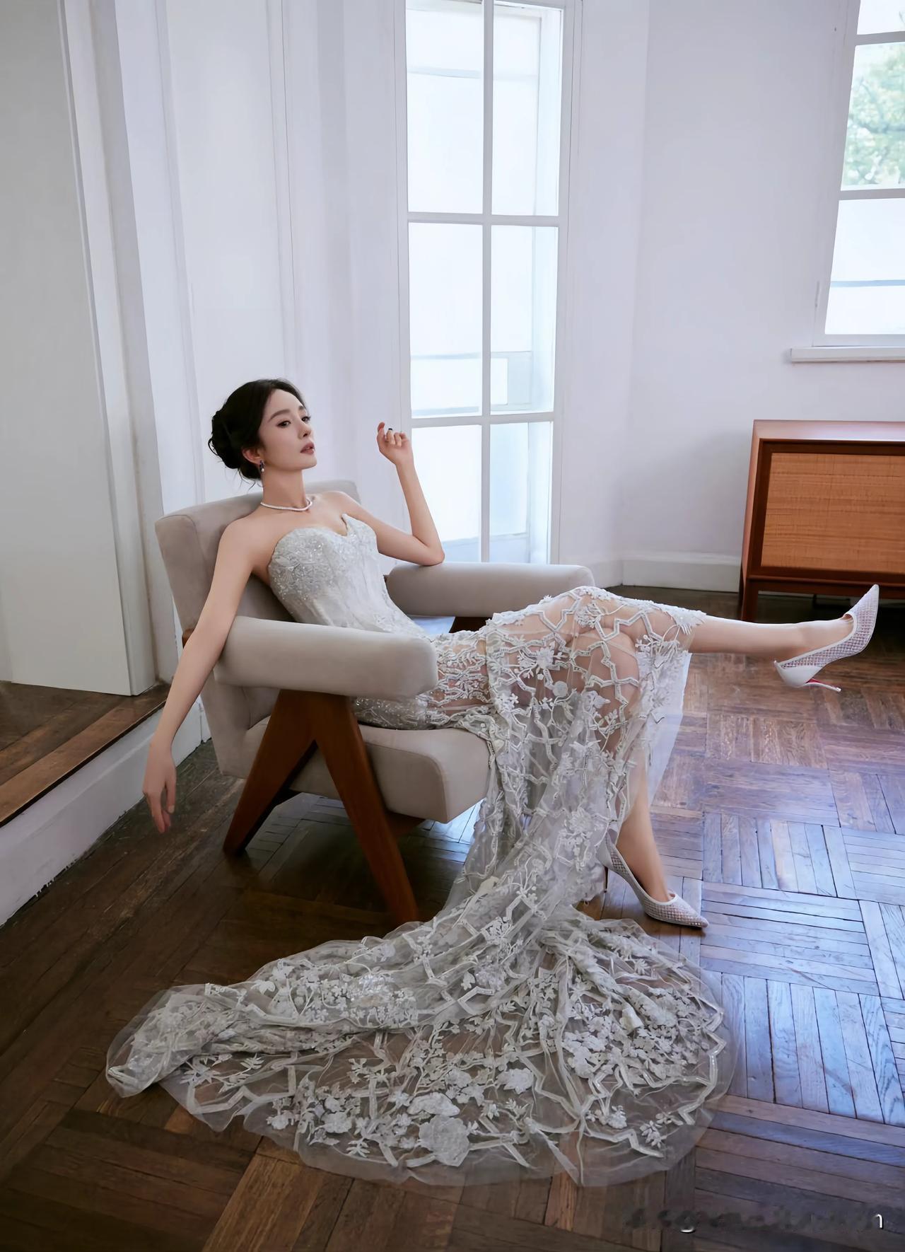 杨幂身着一袭精致的抹胸蕾丝长裙，慵懒地倚坐于椅上，优雅与性感并存。蓬松的裙摆似绽