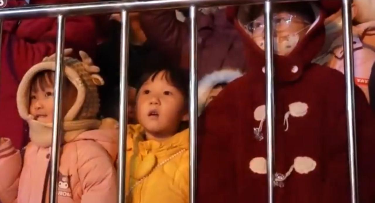 夜色里的城市街巷满是热闹气息，孩童裹着厚实的棉服，扒着栏杆睁大眼睛望向远方，脸上