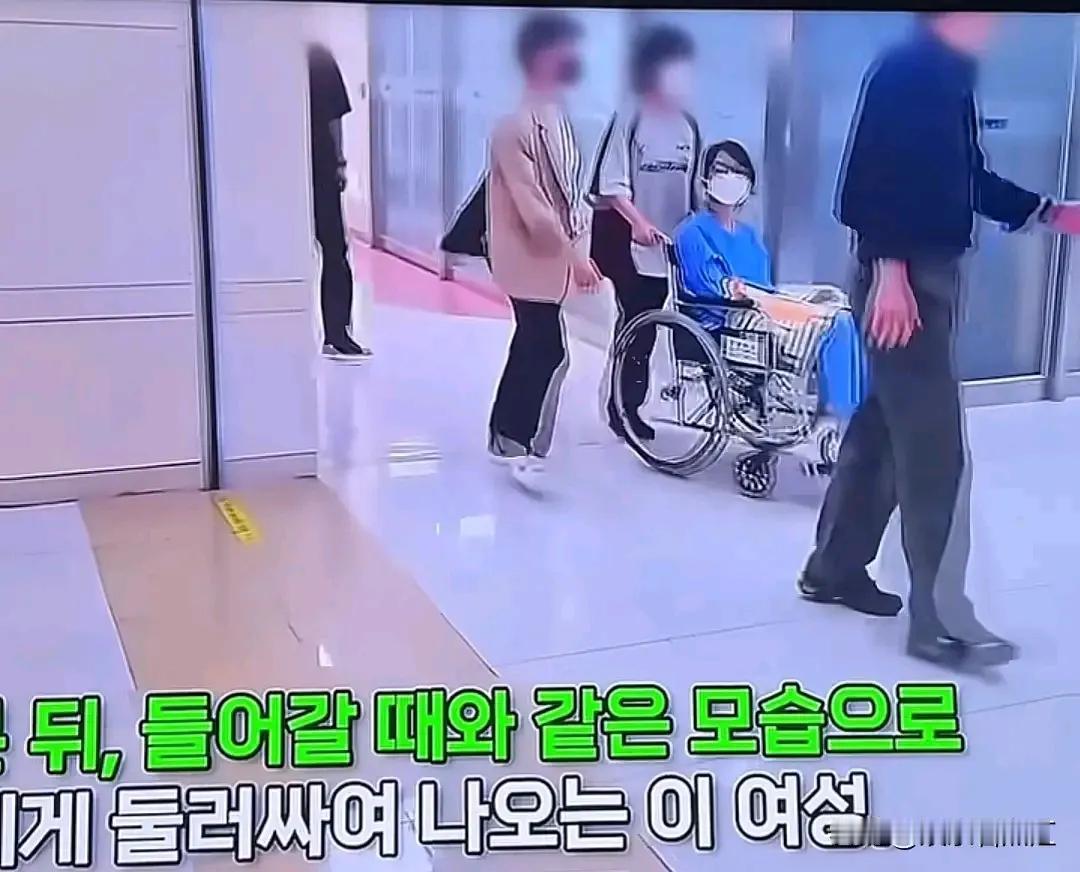 韩国前第一夫人在被拘留后首次露面，被拍到穿着病号服坐着轮椅就医。
画面中金建希双