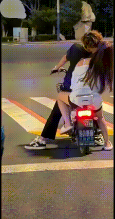 十字路口等红灯，一对小情侣骑电动车停车等候，双脚撑地稳住车子。小伙子忍不住转头和