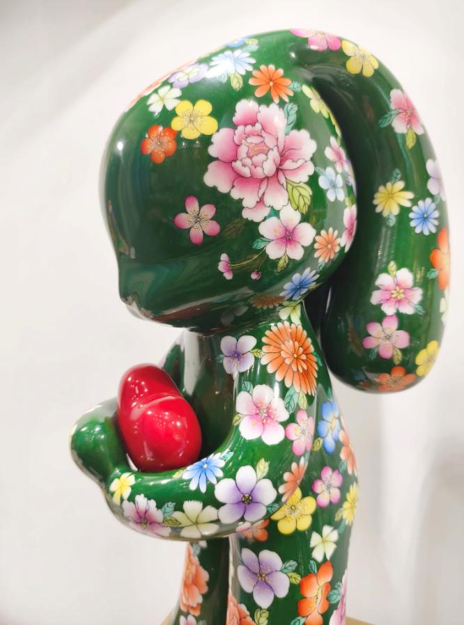 深绿为底，繁花满衣，潮玩形态的雕塑怀抱一颗红心，以传统花卉纹样碰撞当代艺术语言