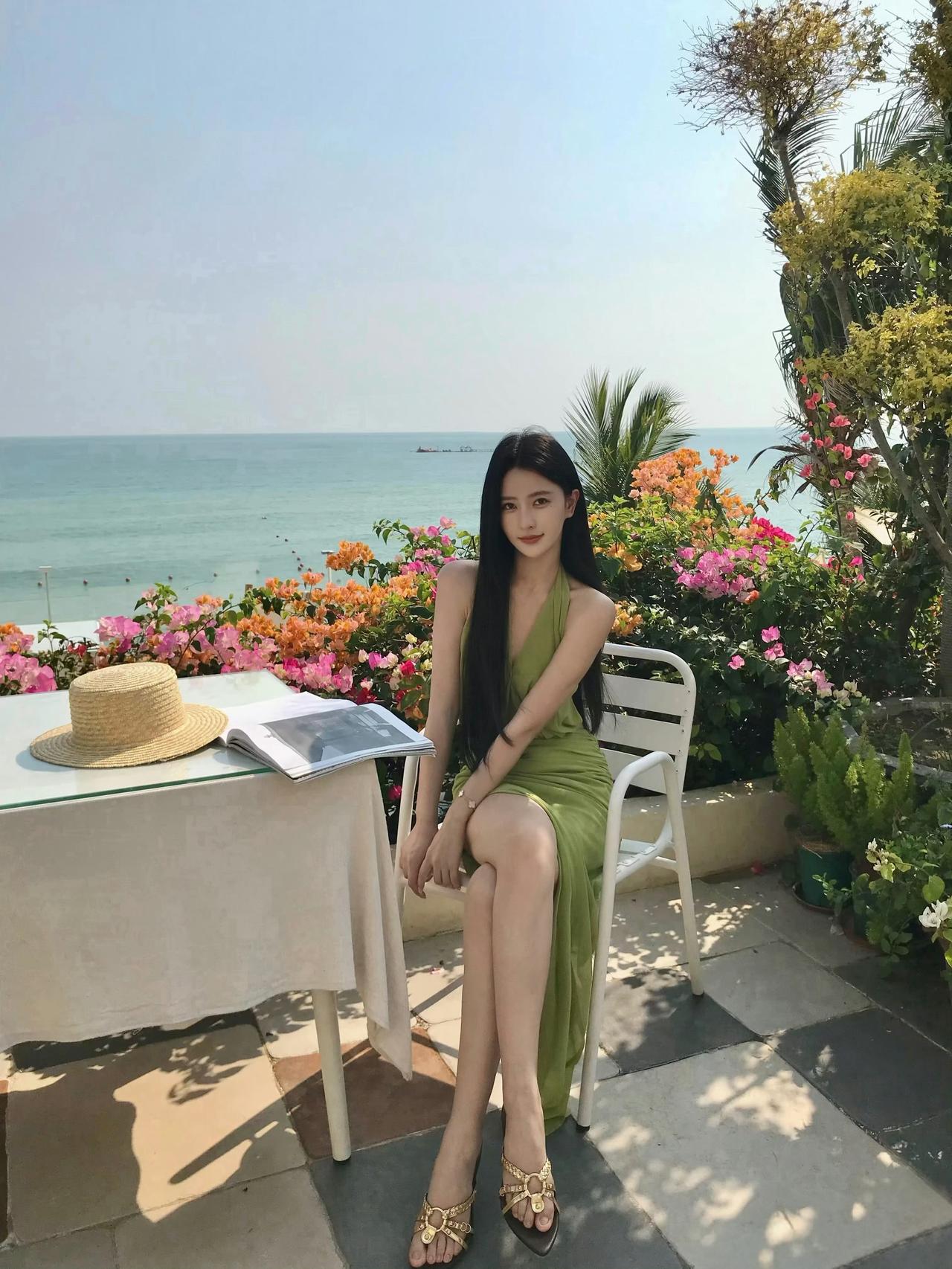 绿色吊带开叉长裙，搭配金色凉鞋，周围繁花盛开，海景迷人，整体氛围清新惬意，尽显悠