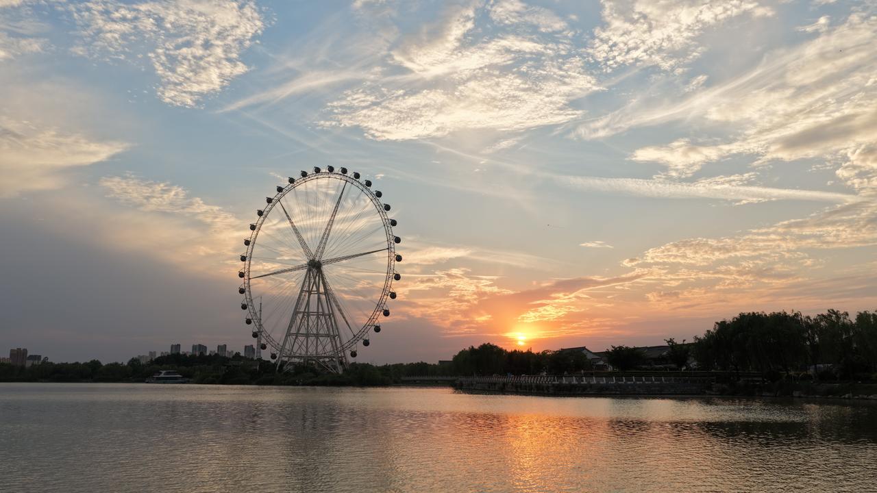 晚风拂过堤岸垂柳，古建白墙黛瓦错落排布，河道蜿蜒穿城，远处楼宇与风车隐在薄暮里。