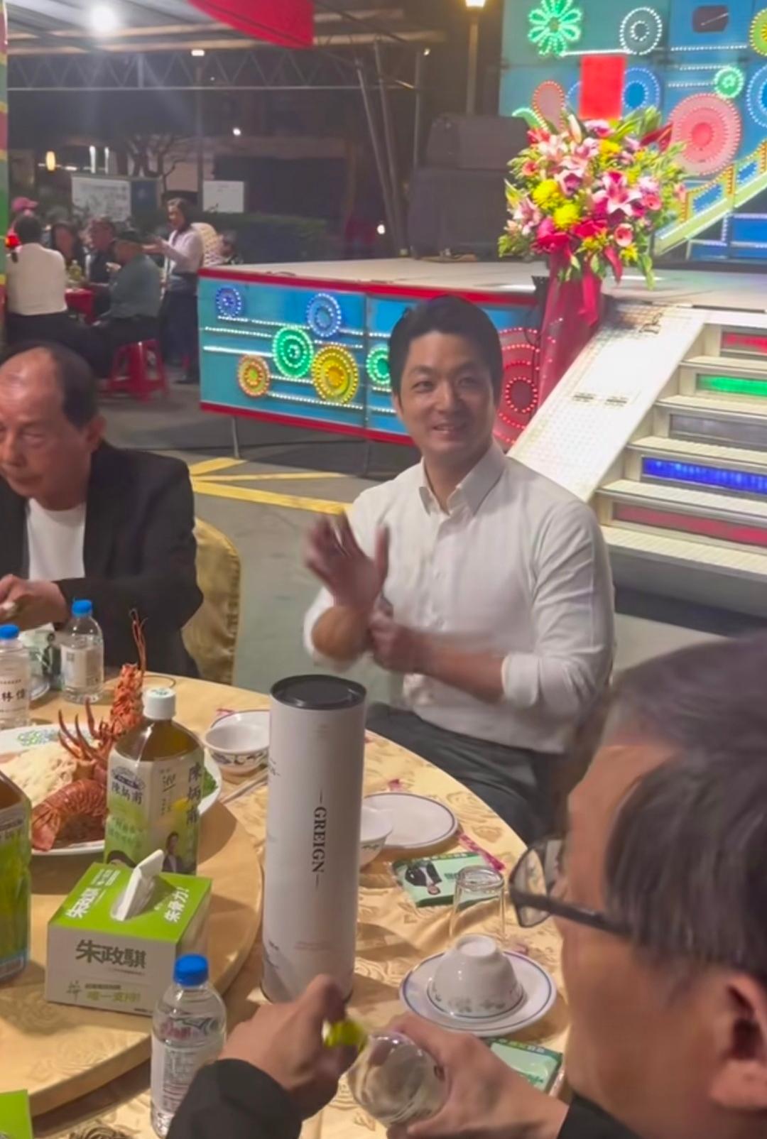 蒋万安像是参加农村大席，旁边有一个临时搭建的舞台，还有一堵用塑料布隔的挡风墙，远