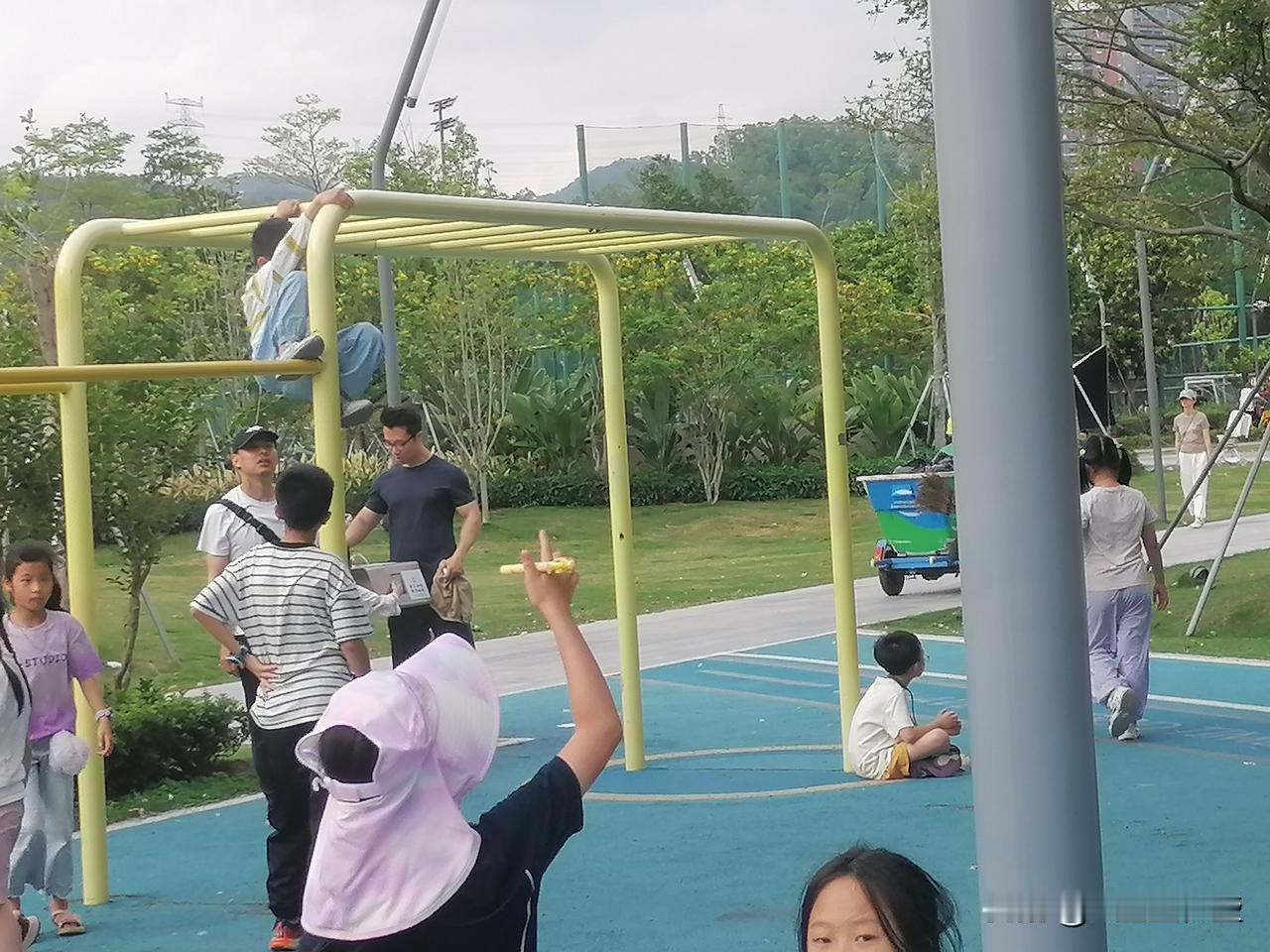 小孩练杠杠，大人下边护着，一女士手指着天上，看什么呢？
