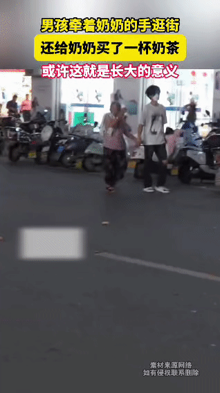 在一条热闹的街巷里，一个男孩紧紧牵着奶奶的手，奶奶握着男孩买的奶茶，小口啜饮着，