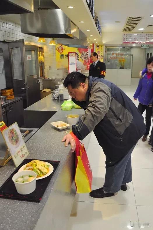 某餐厅没有座位了，一位老吃客在销售台边上，将就着站立用餐。也许是实在饿得慌或者急