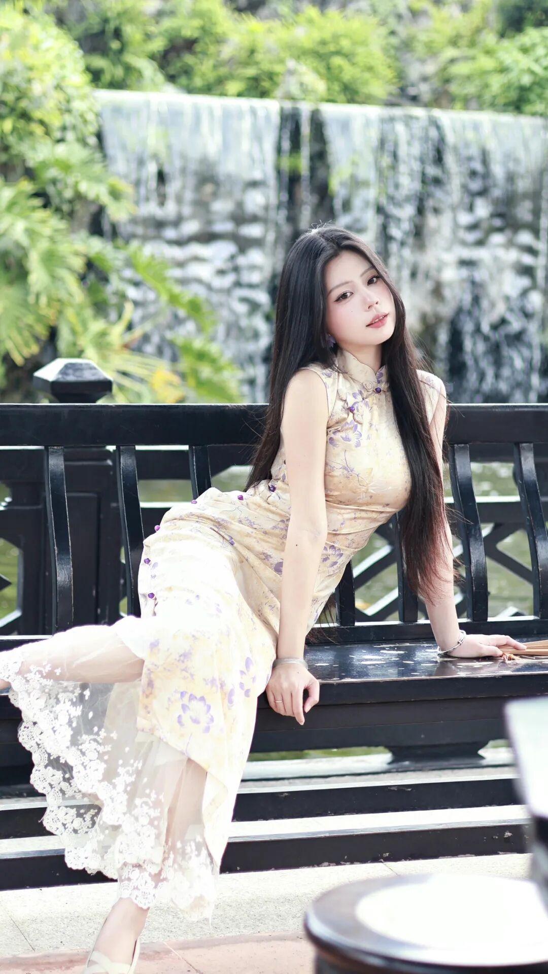 旗袍以温润的米杏色为底，缀着淡雅的紫色碎花，无袖设计勾勒出优美的肩颈线条，裙摆处