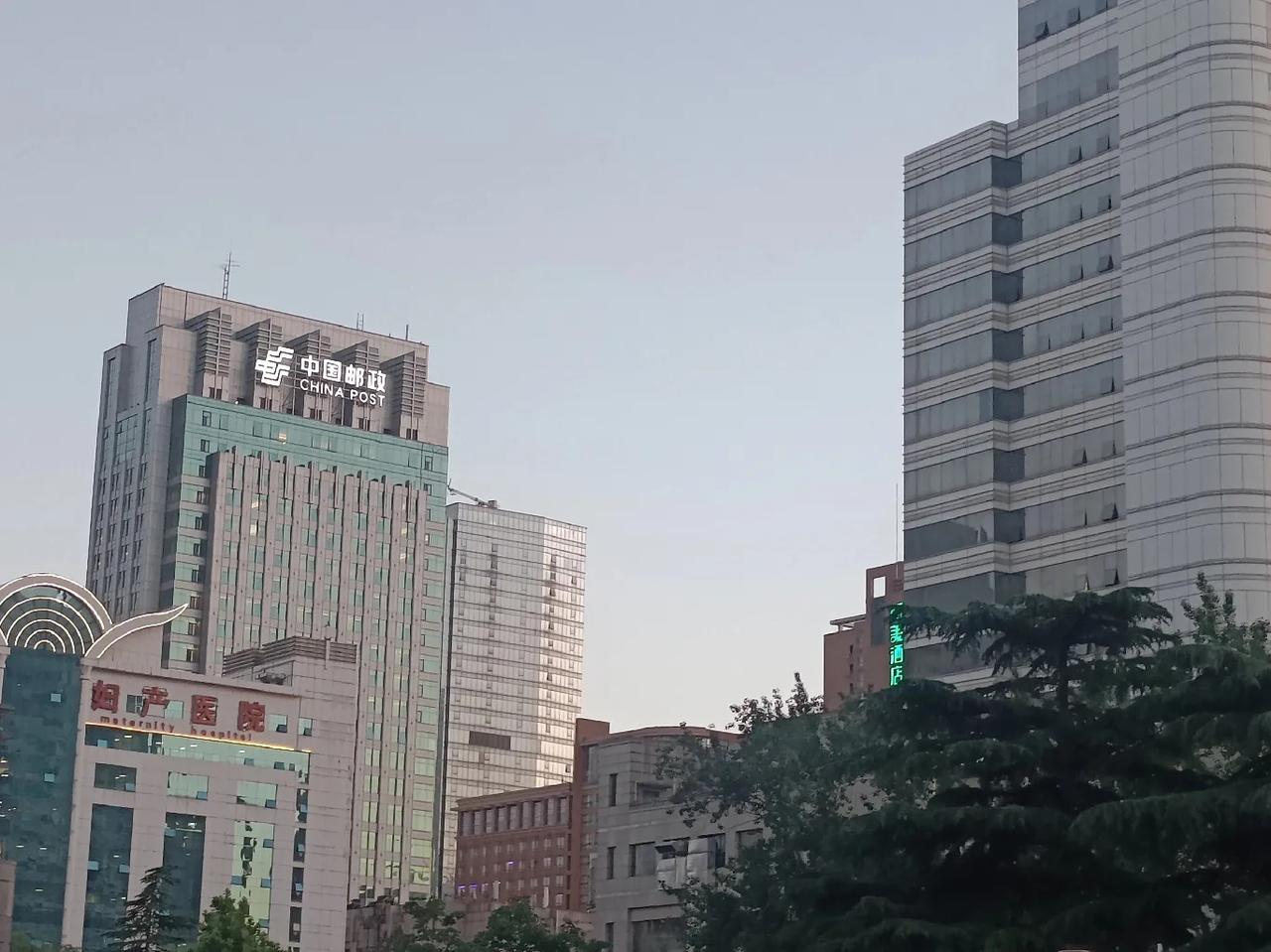 夕阳缓缓沉落楼宇之间，给林立的高楼镀上一层暖橙柔光。街道车流渐密，车灯汇成蜿蜒流