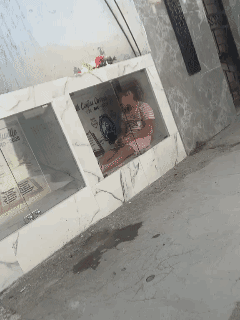 在厄瓜多尔的墓园里，女孩坐在母亲的墓碑边，手里把玩着小玩具，玻璃映着她的笑脸。或