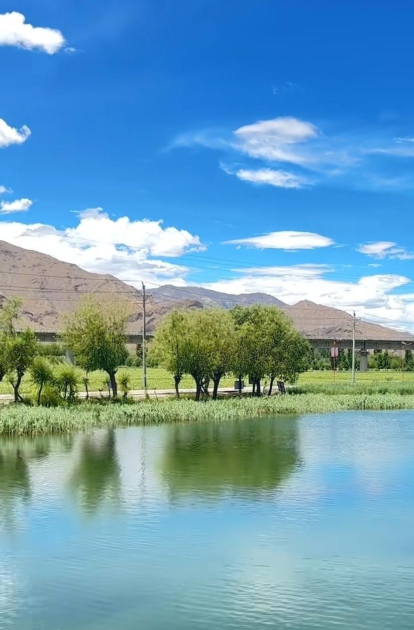 宁静的湖面倒映着蓝天白云，远处的山峦和绿树构成了一幅美丽的自然画卷。阳光洒在湖面