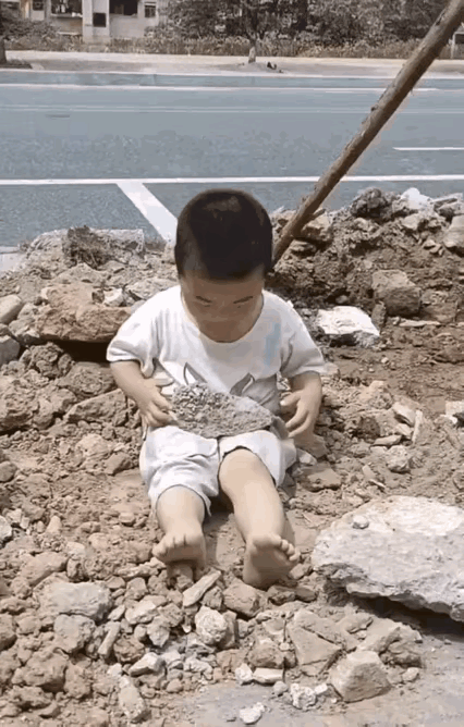小男孩坐在乱石堆里面，双手举起了一块石头，然后往地上砸了下来，砸的时候砸到了自己