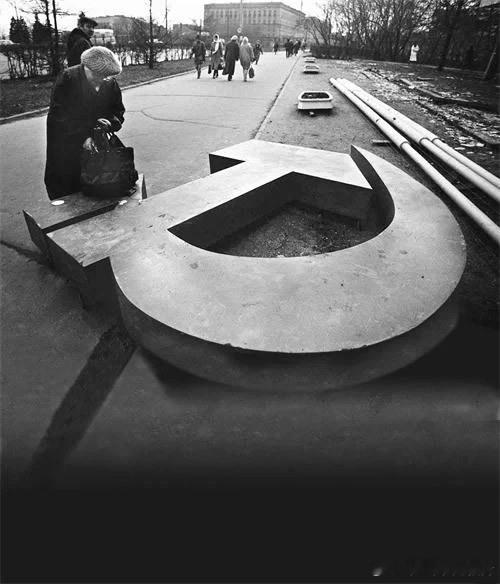 1991年苏联解体，莫斯科街头，一位妇女把她的包放在倒地的镶刀斧头雕塑上掏东西 