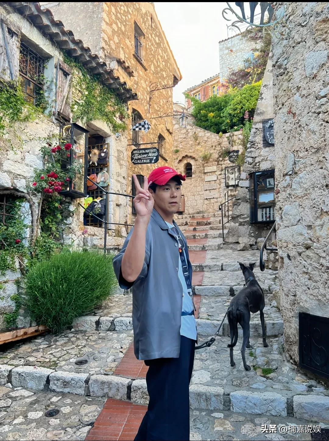 彭于晏现身街头遛狗，戴着红色的棒球帽，穿着一件灰蓝色的风衣配运动裤，肤色偏黑，个