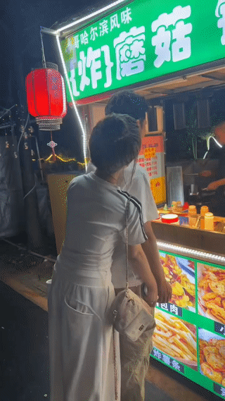 男子正站在一个小吃摊前面购买小吃。
而他的女朋友则默默的站在他身后，看起来非常羞