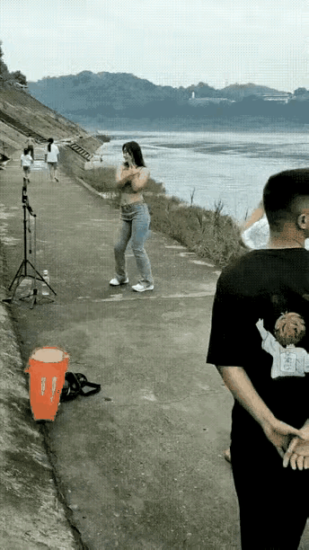 河边步道上，一个女主播正对着手机跳舞直播。有对路人刚好经过，那个穿黑衣服的男子压