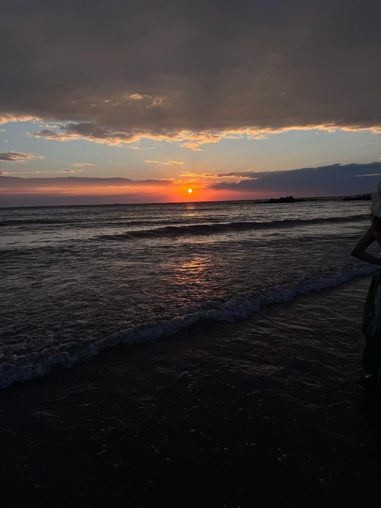 海浪层层漫过沙滩，落日坠入海面，把细碎金光铺在起伏的波涛之上。
云层被落日染出温