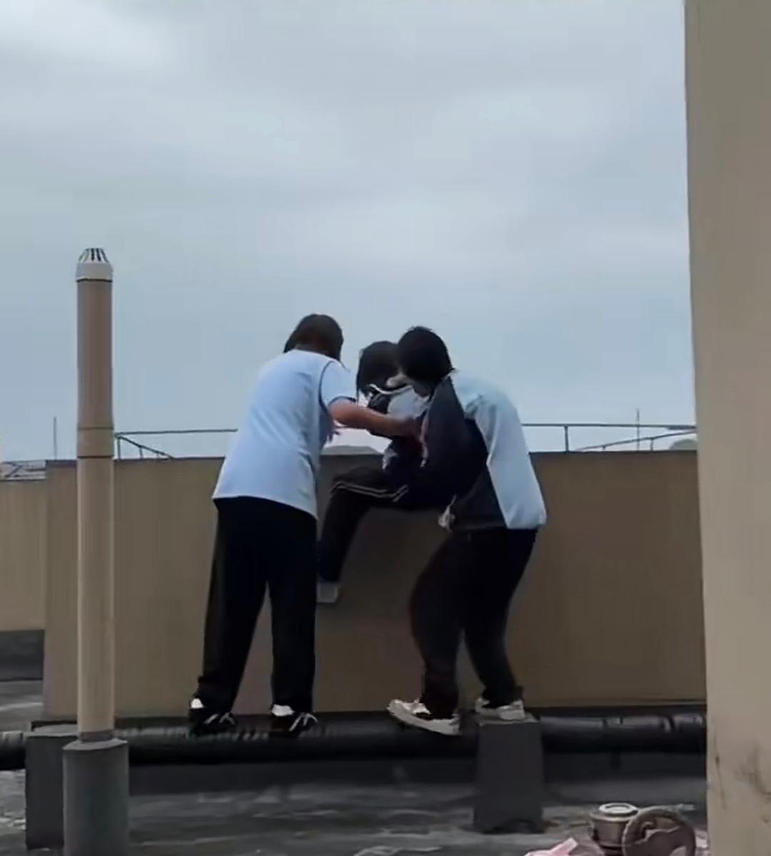 高楼天台，两个女生架着另外一名女孩往护栏外面抬，半个身子已悬在几十米高空。楼下业