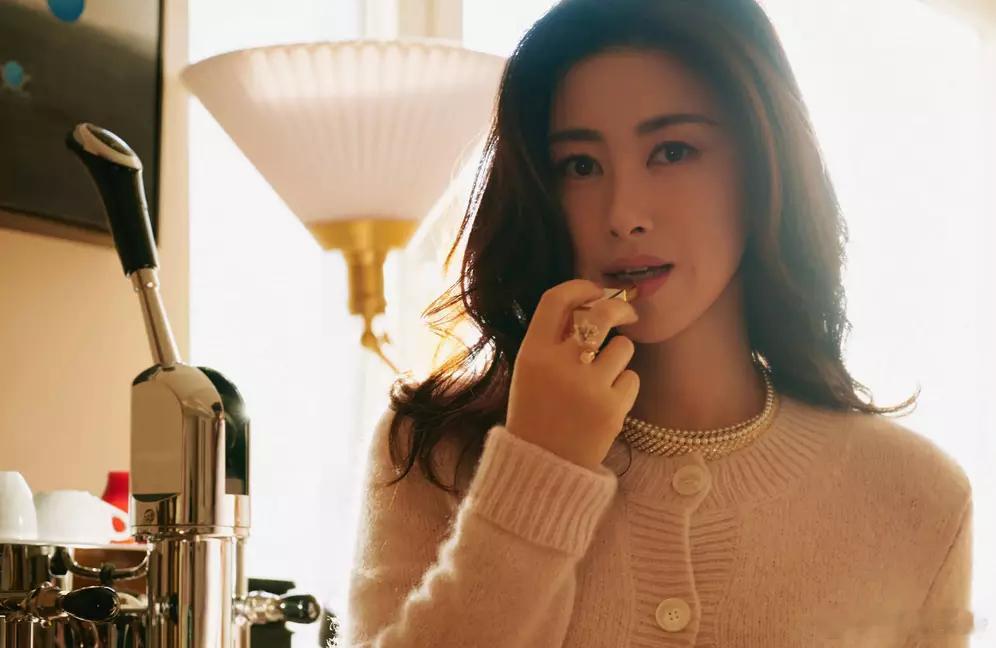 朱珠妆容精致，身着粉色针织开衫，佩戴珍珠项链与戒指，在暖光下涂口红，尽显优雅知性